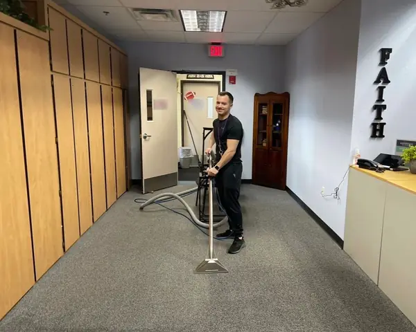 A crew member in black smiles at the camera as he runs a carpet cleaning wand over gray carpet in a front office lined with wooden cabinets