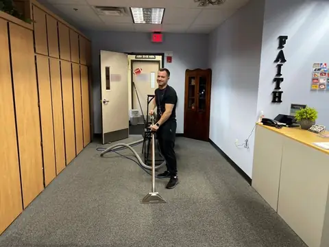 A crew member running a carpet cleaning wand over gray carpet in a front office, with the hoses on the floor behind him