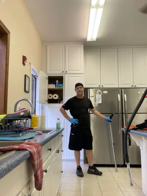 A crew member in black with blue gloves standing in a kitchen with white cabinets and two steel refrigerators, holding a vacuum wand