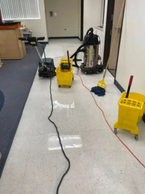 A rotary floor machine, a steel wet vacuum, two yellow mop buckets and a mop on a tile floor, with extension cords running toward the camera
