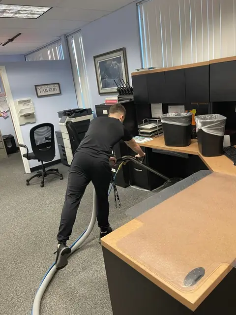 Gary bending to run a carpet cleaning wand under a reception desk in an office, with the hose trailing across the carpet