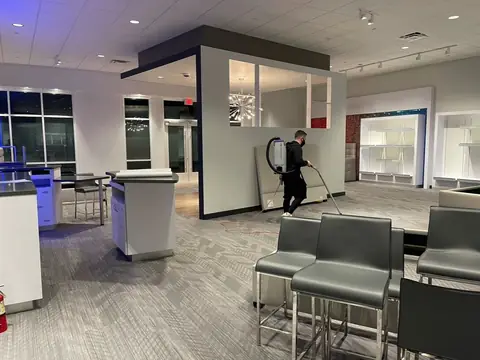 Gary wearing a backpack vacuum, working across the carpet of a showroom at night, with gray chairs in the foreground