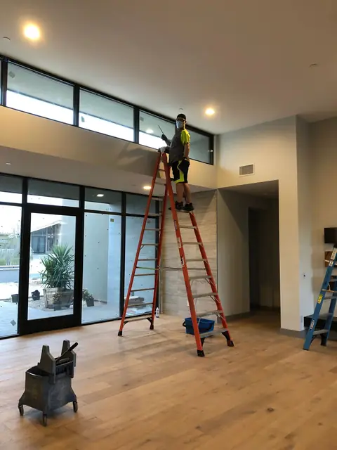 James on a tall orange ladder at the high windows of a room with a wood floor, a mop bucket in the foreground