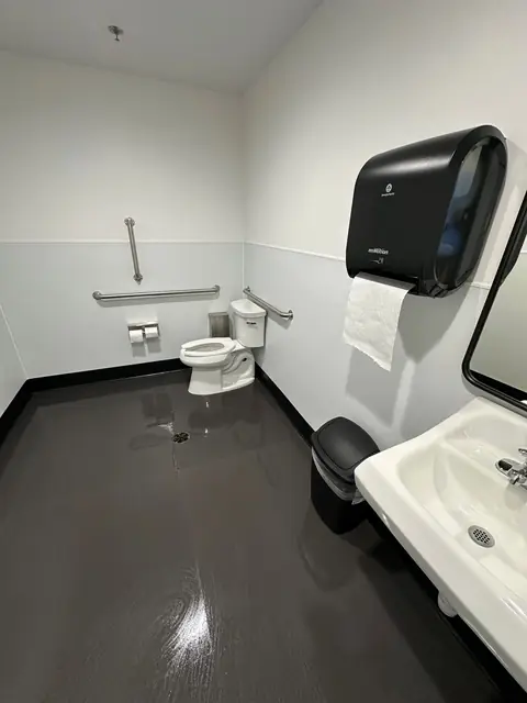 An accessible restroom with a glossy dark floor, a toilet with grab bars, a paper towel dispenser and a sink in the foreground
