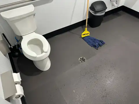 A toilet, a string mop leaning against the wall beside a trash bin, and a floor drain in a dark floor
