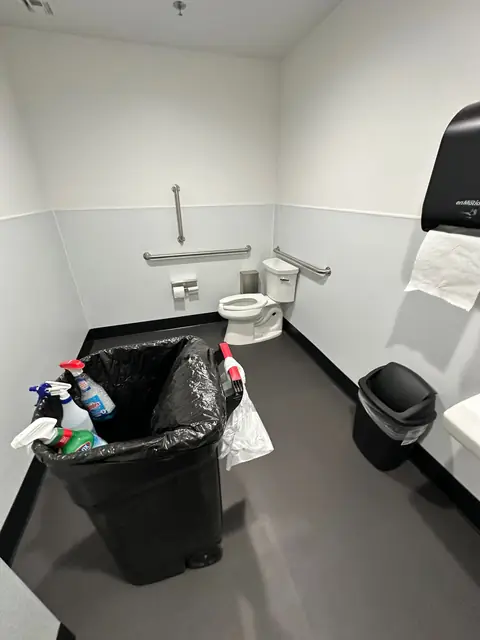 A restroom with a toilet and grab bars, a small trash bin, and spray bottles standing on the rim of a large lined trash bin