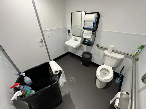 A restroom with a wall sink, a toilet with grab bars, a paper towel dispenser and a floor drain, and spray bottles in a lined trash bin in the foreground