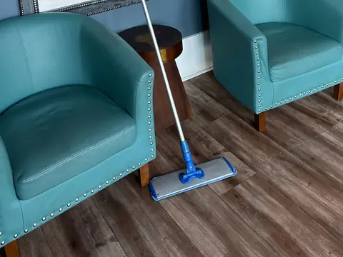 A flat mop leaning between two teal armchairs on a wood-look floor