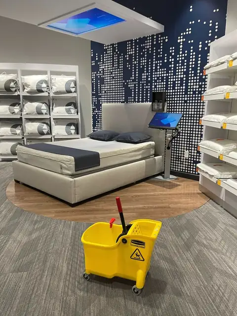 A yellow mop bucket on the carpet of a mattress showroom, in front of a bed on display and shelves of pillows